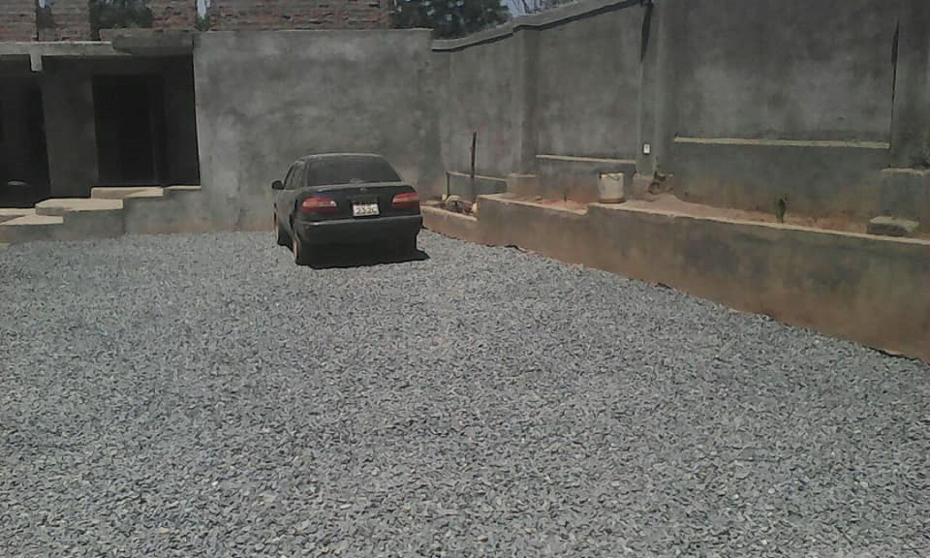 Un coche aparcado en un área de grava con un muro de hormigón y escaleras que suben a la entrada de un edificio.