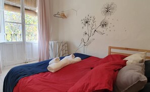 A hotel room with a bed, a radiator, a window with blinds, and a wall with a floral mural.