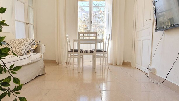 Une pièce avec un canapé blanc, une table à manger avec des chaises, un téléviseur fixé au mur et une fenêtre avec des rideaux.