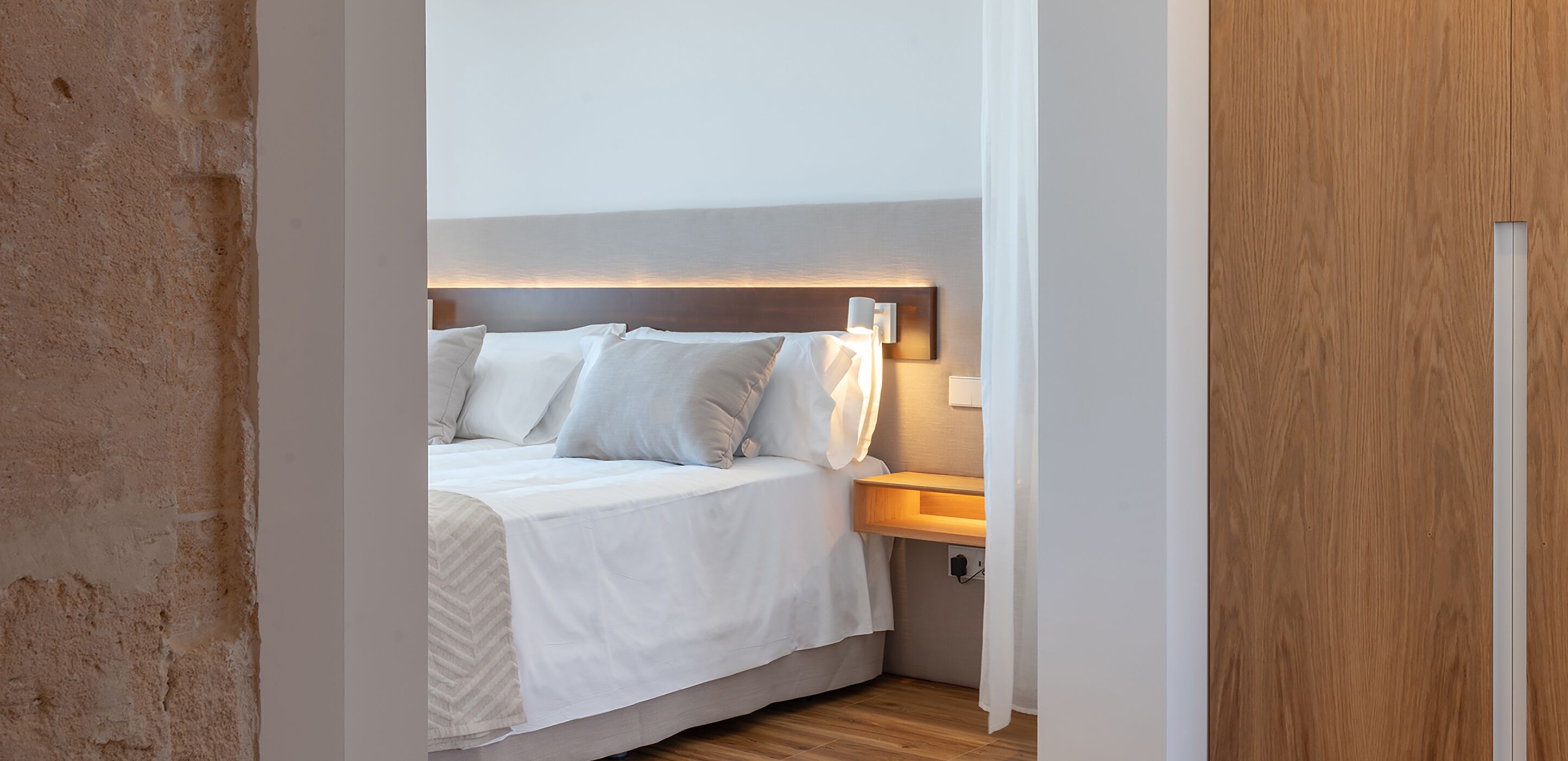 A neatly made bed with white linens and a headboard, a bedside table with a lamp, and a wooden floor.