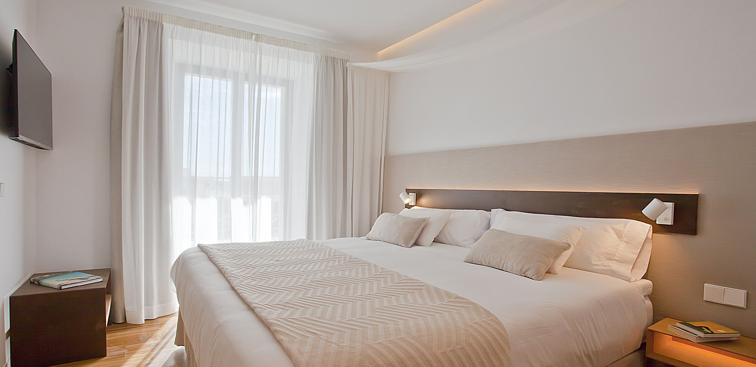 Una habitación de hotel moderna con una cama grande, un televisor de pantalla plana en la pared y una mesita de noche con lámpara.