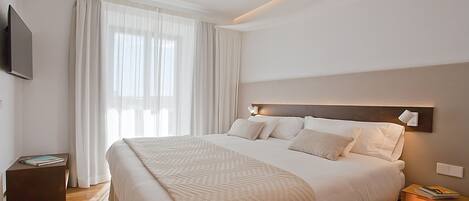 A modern hotel room with a large bed, a flat-screen TV mounted on the wall, and a bedside table with a lamp.
