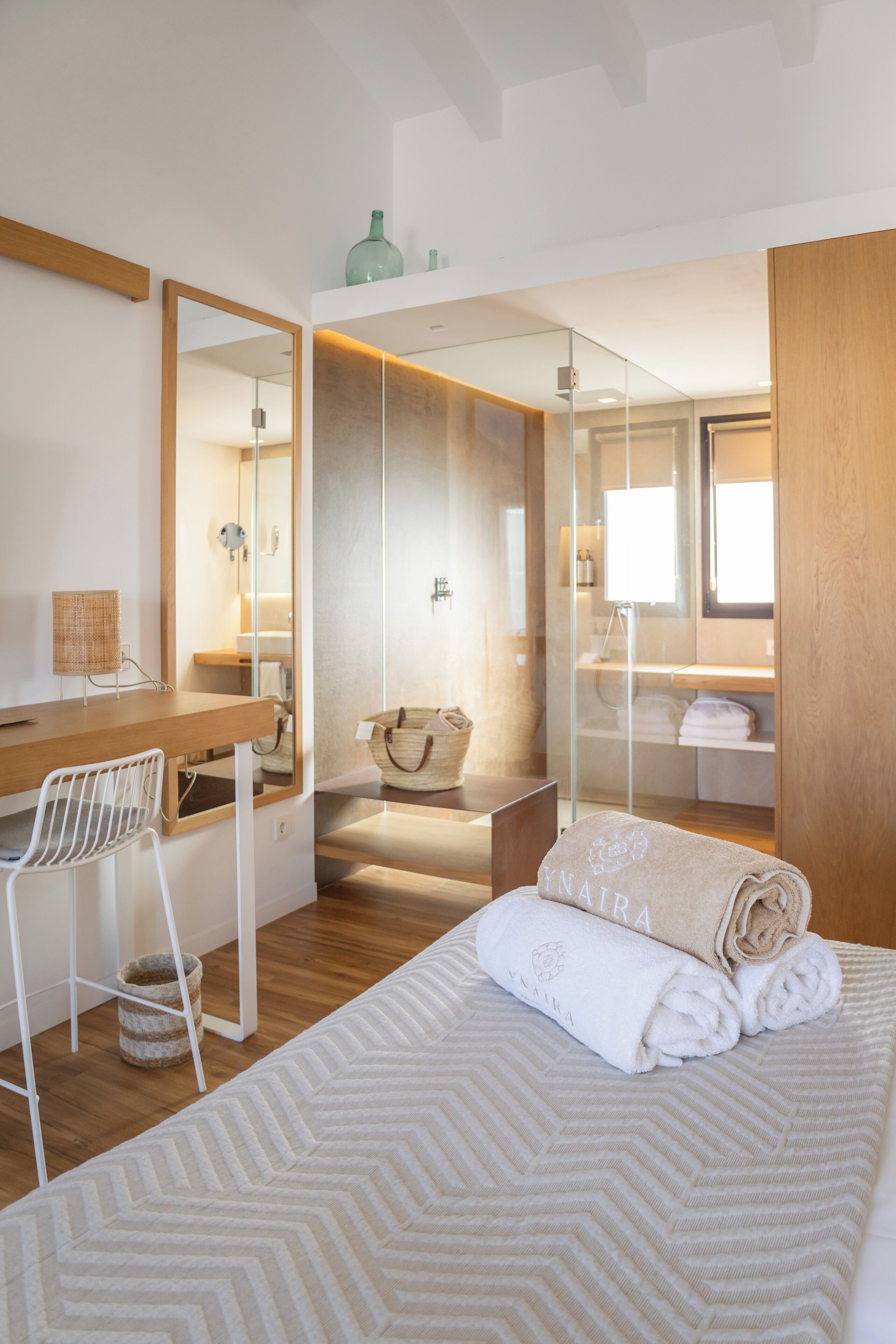 Une chambre moderne comprenant un lit, un bureau, une chaise, un miroir et une salle de bain visible à travers une cloison en verre.