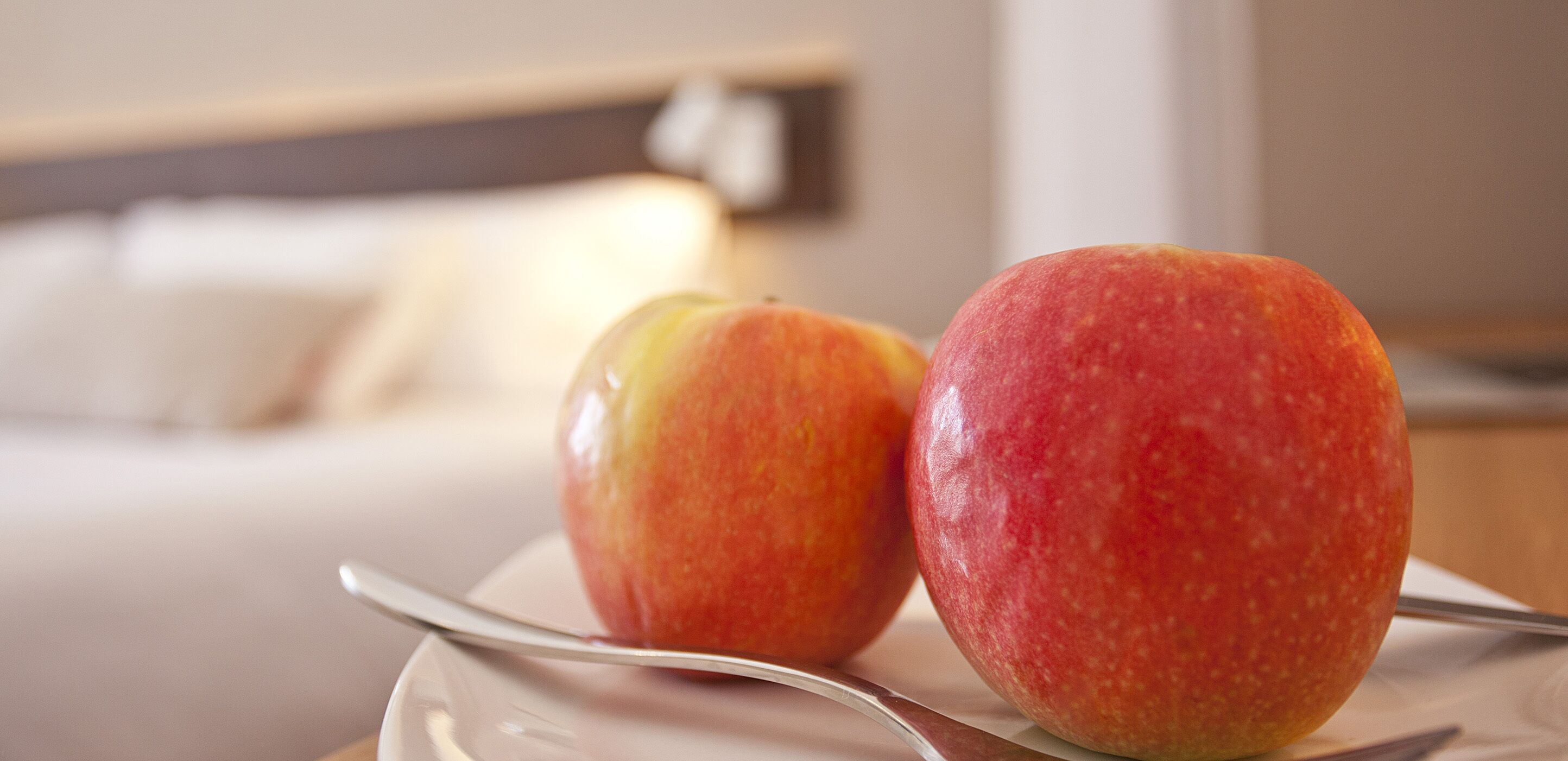 Two apples on a plate with a spoon, in a room with a bed and a nightstand.