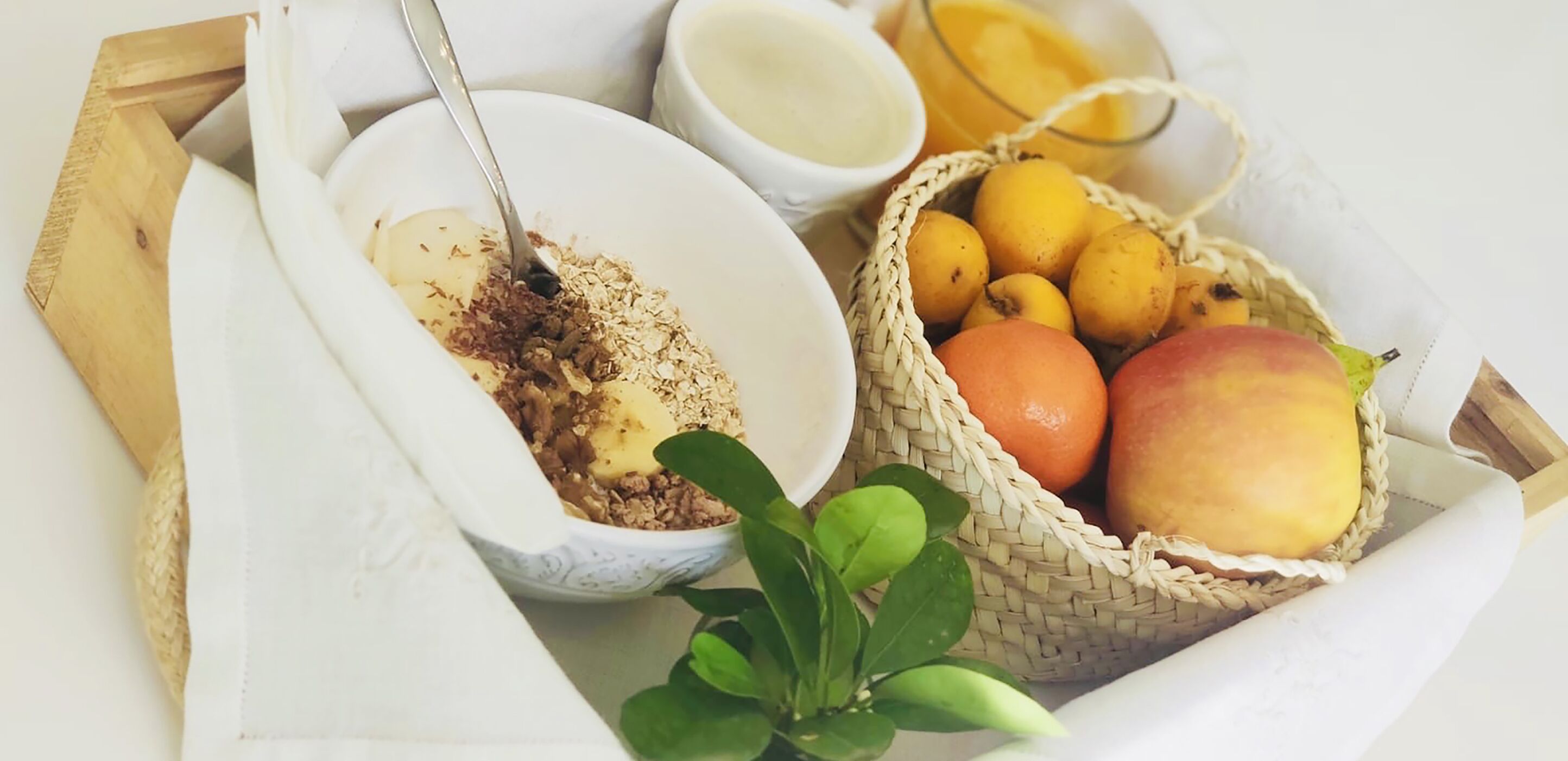 A breakfast setting with a bowl of cereal, a basket of fruits, and a cup of milk.