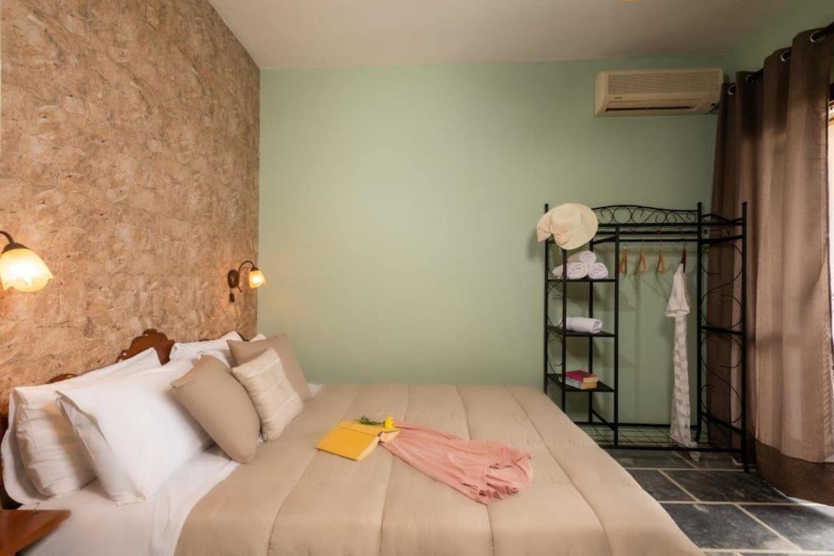A bedroom with a bed, pillows, a bedside table, a wall-mounted shelf with towels, and an air conditioning unit.