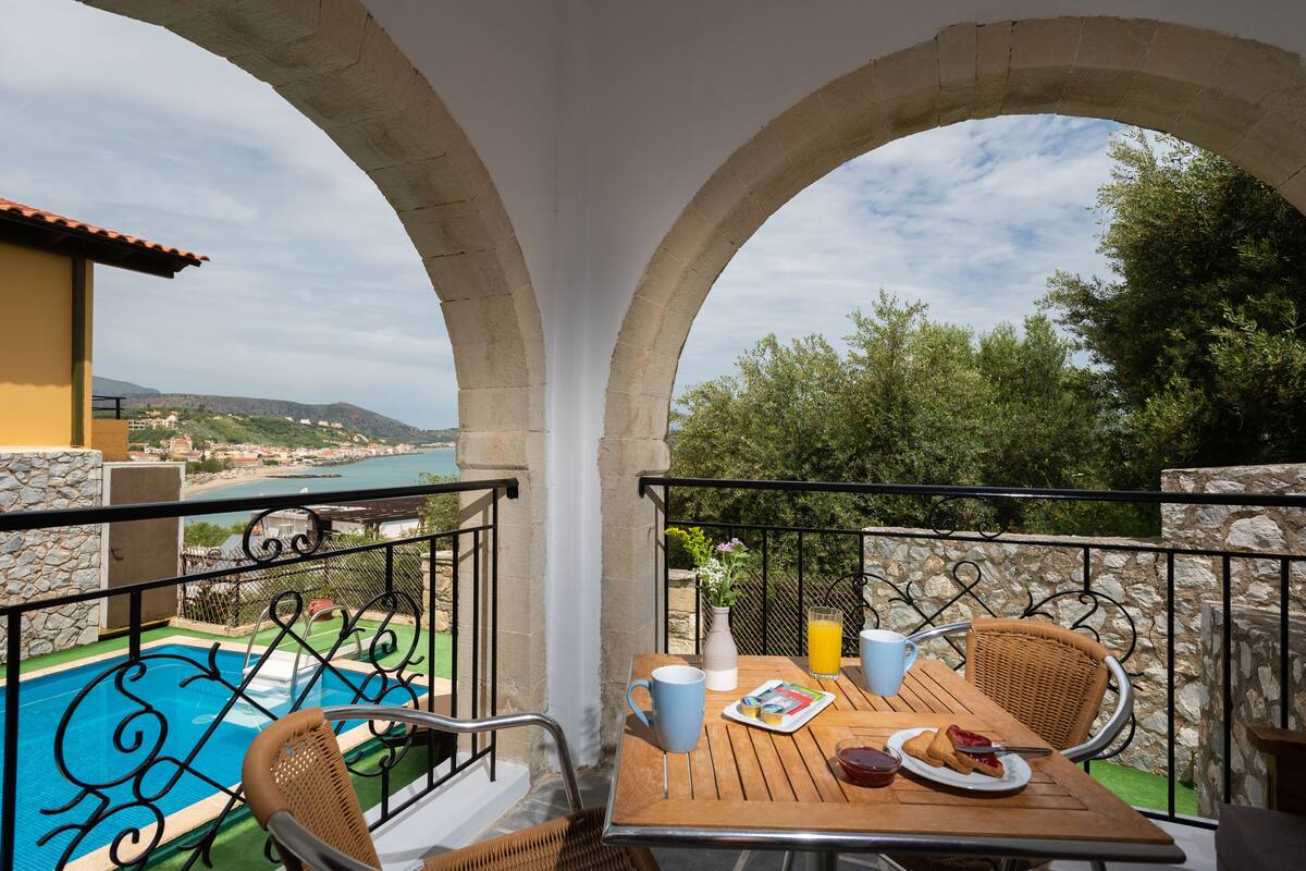 A balcony with a table set for breakfast, overlooking a swimming pool and a scenic view of the sea and hills.