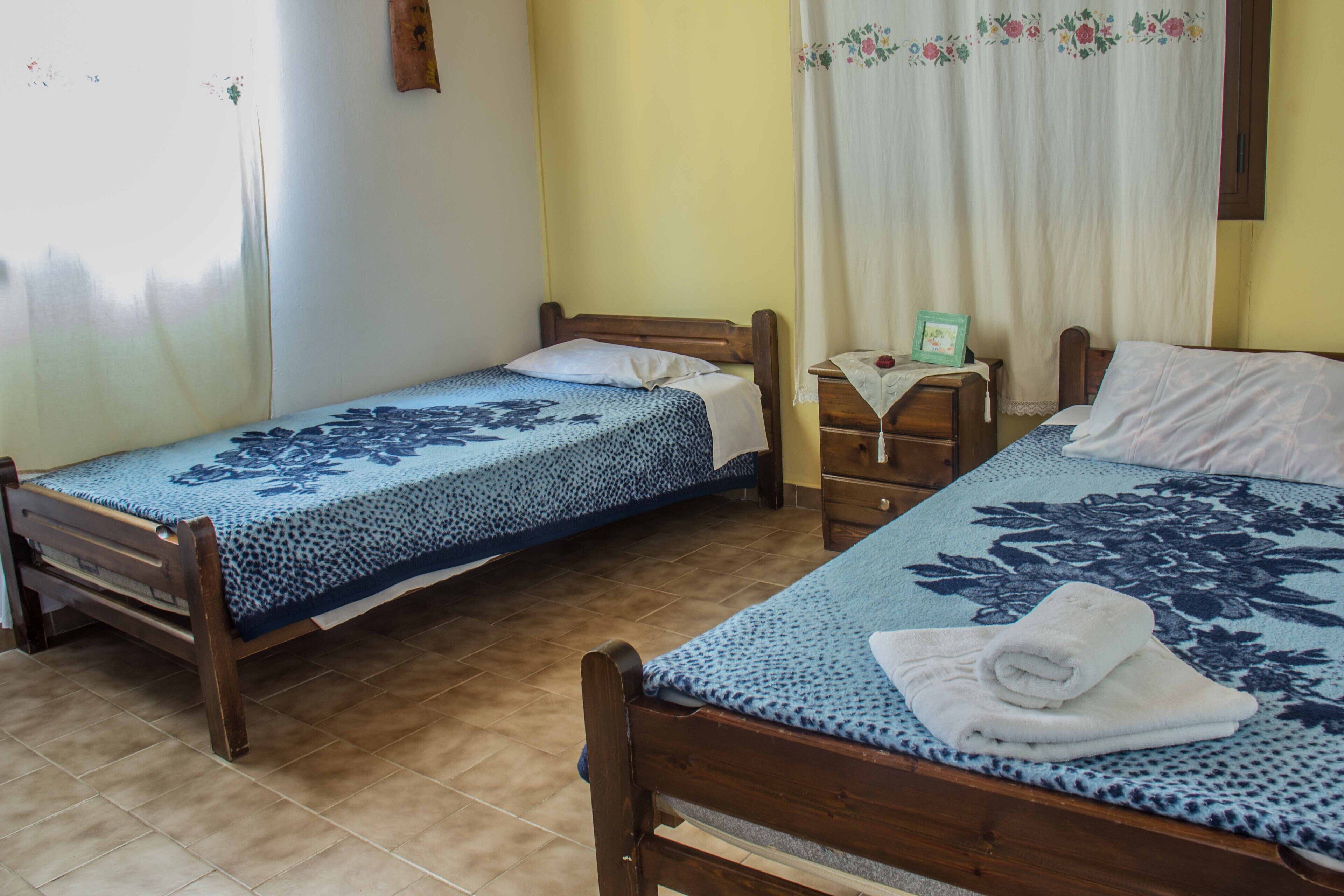 Une chambre avec deux lits en bois, chacun recouvert d’une couvre-lit à motifs floraux. Entre les lits, on trouve une petite table de chevet en bois, sur laquelle se trouve une lampe et un tableau encadré.