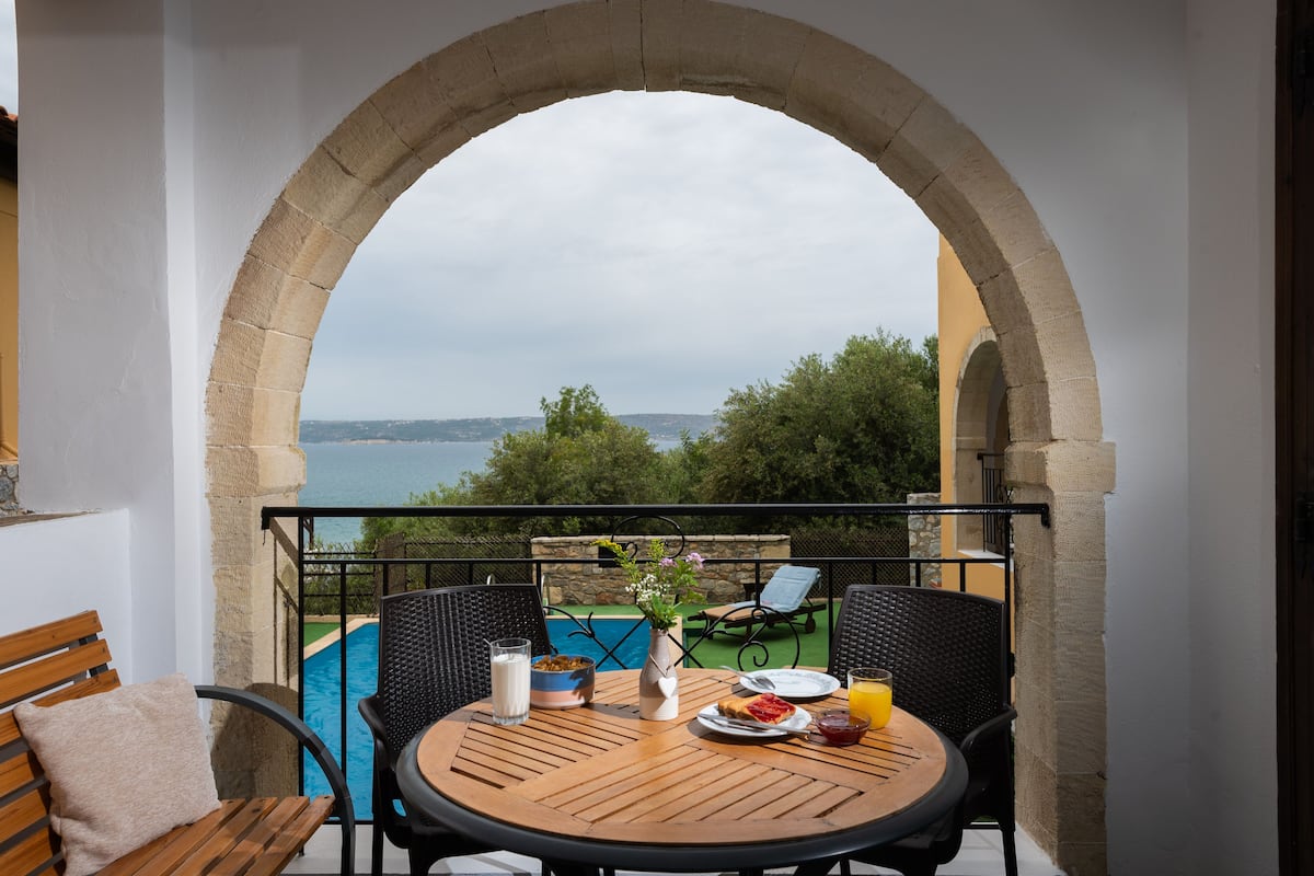 A balcony with a view of the sea, a round wooden table with breakfast items, and a stone archway.
