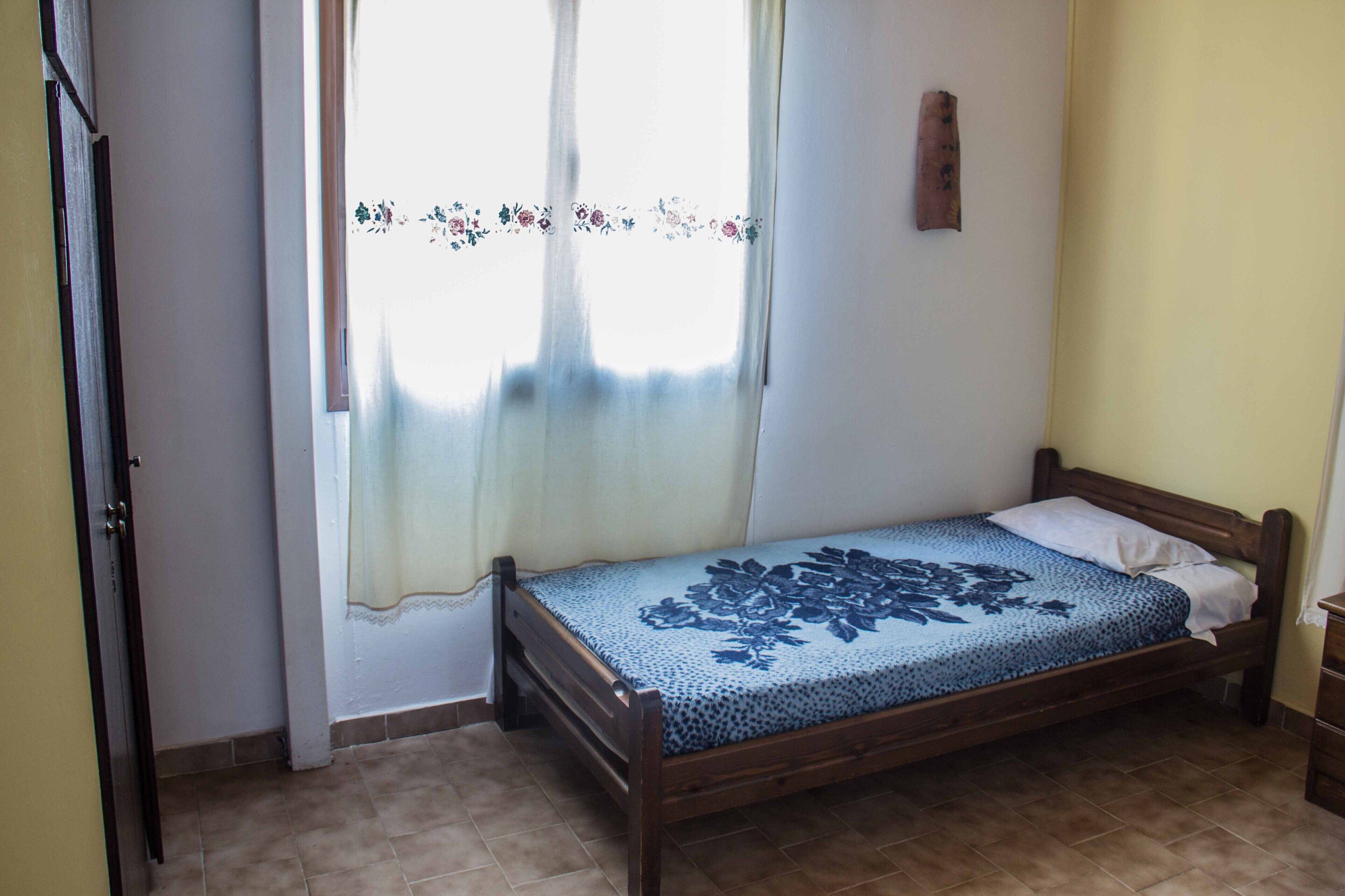 Una habitación pequeña con una cama individual, un marco de madera, una colcha de motivos florales y una ventana con cortinas de encaje.