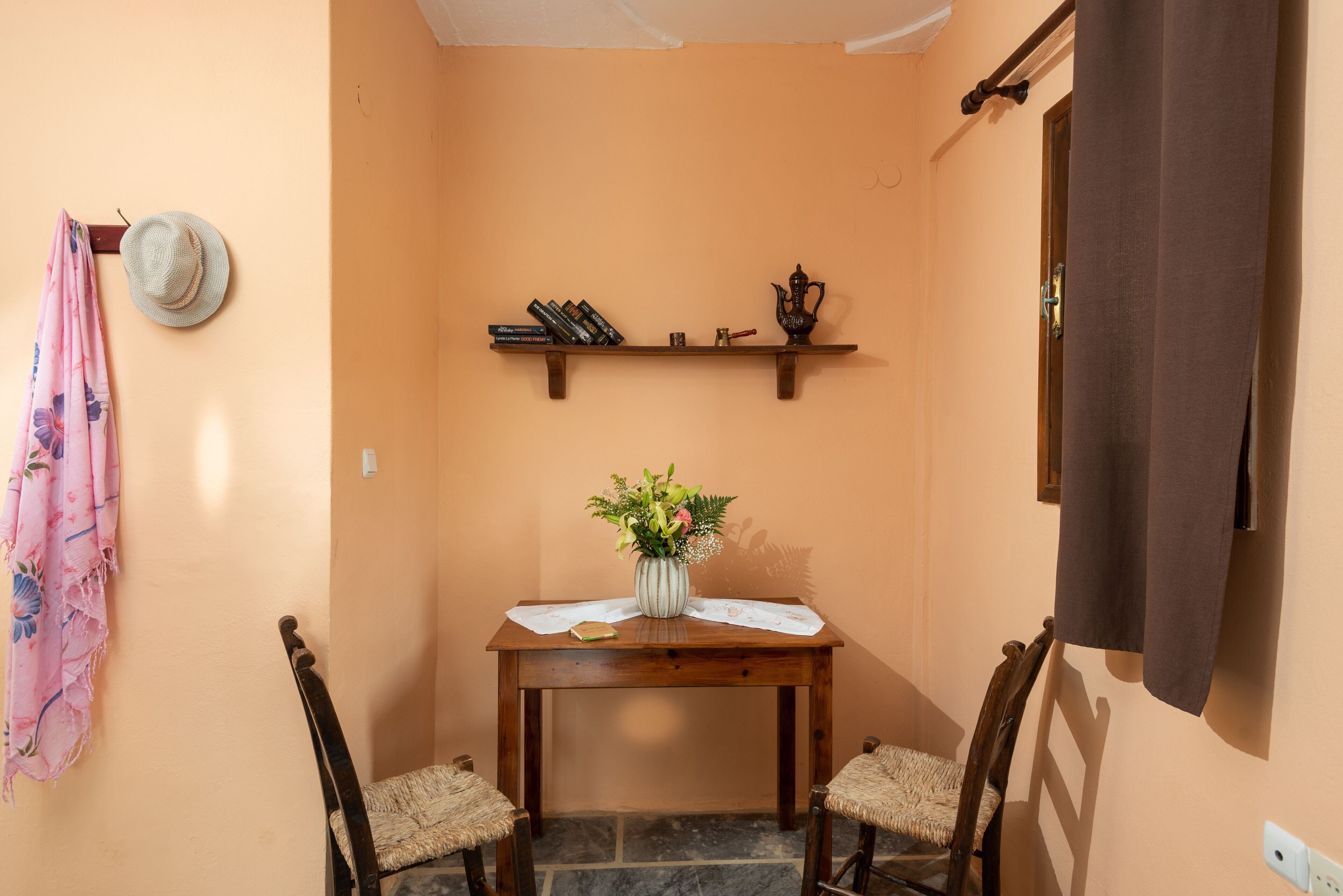 A small dining area with a table, chairs, a hat on the wall, a shelf with books and a teapot, and a window with a curtain.