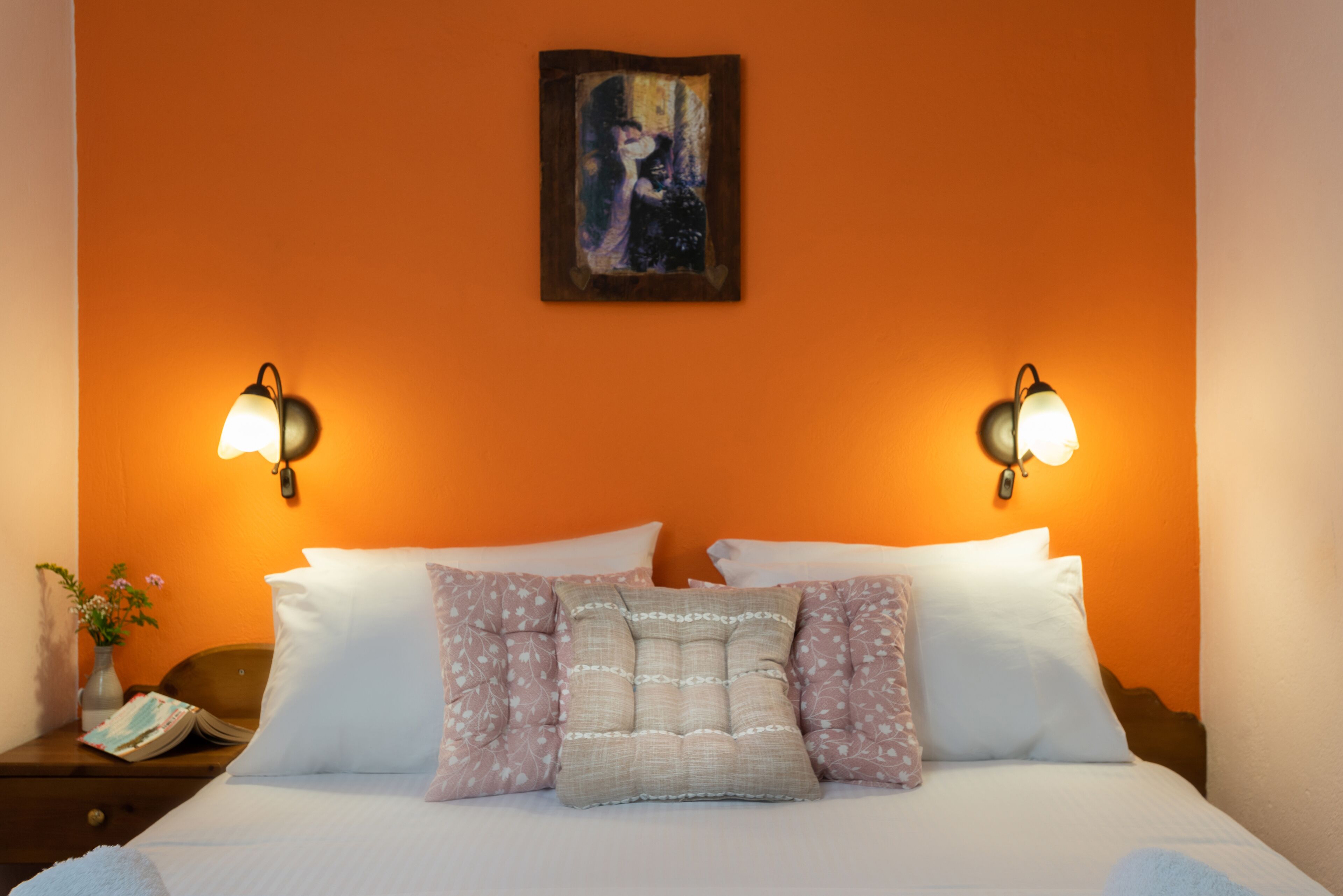 A hotel room with a bed, two wall-mounted lamps, a painting on the wall, and a nightstand with a vase of flowers.