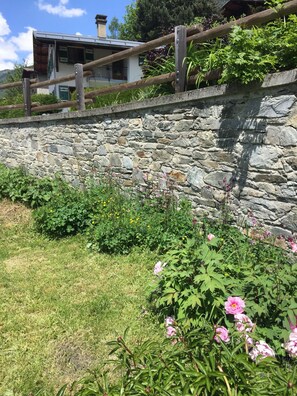 Un muro de piedra con valla de madera, un área verde con flores rosas y una casa al fondo.