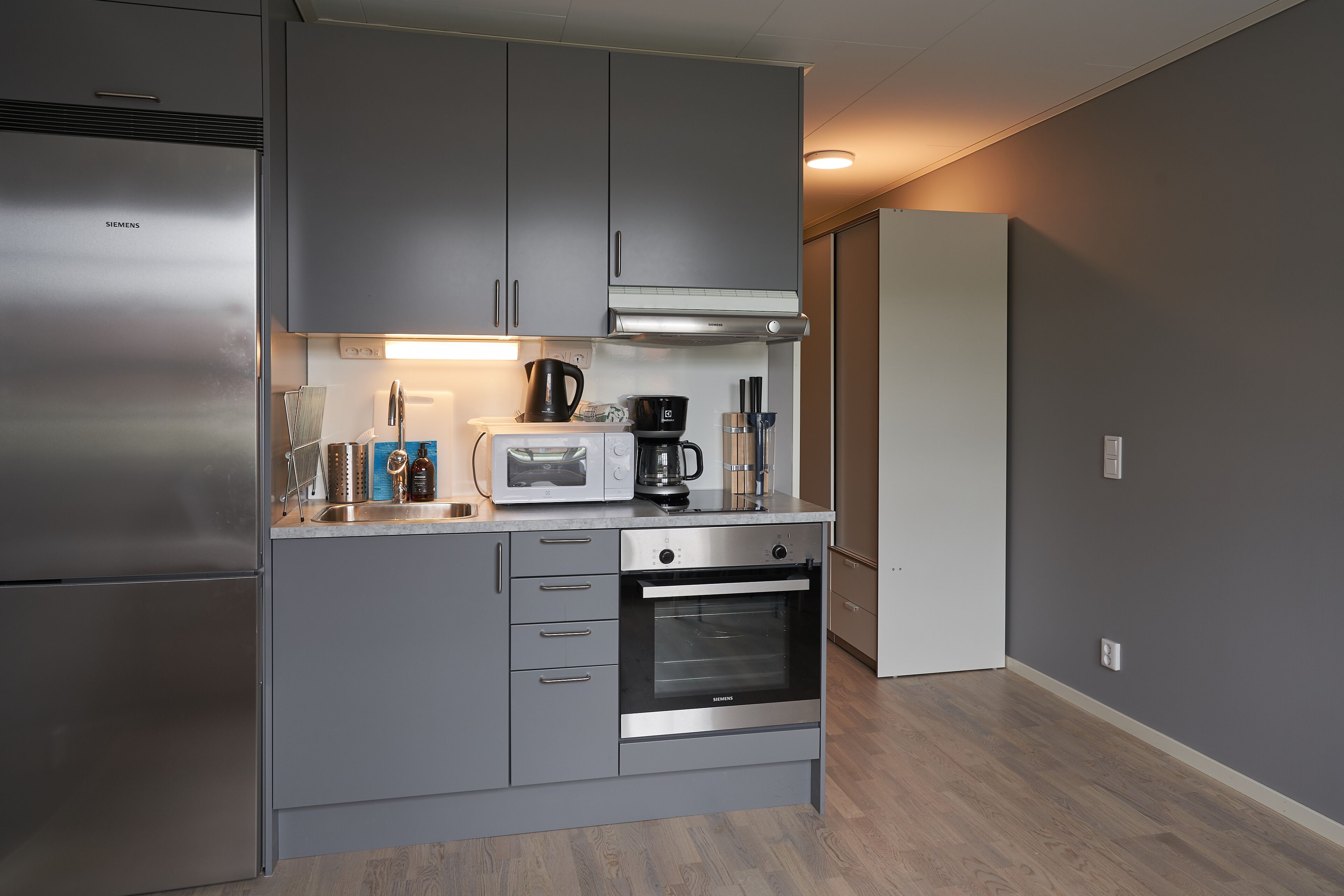 A modern kitchen with grey cabinetry, a stainless steel refrigerator, a microwave, a coffee maker, and an oven.