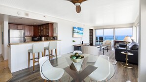 A modern kitchen with wooden cabinets, a stainless steel refrigerator, and a dining area with a glass table and white chairs. - Amazing Sunset Views, Renovated & Furnished Condo at Ilikai Hotel! (Honolulu)
