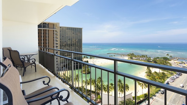 A balcony with a view of a beach, a high-rise building, and a parking area. - Amazing Sunset Views, Renovated & Furnished Condo at Ilikai Hotel! (Honolulu)