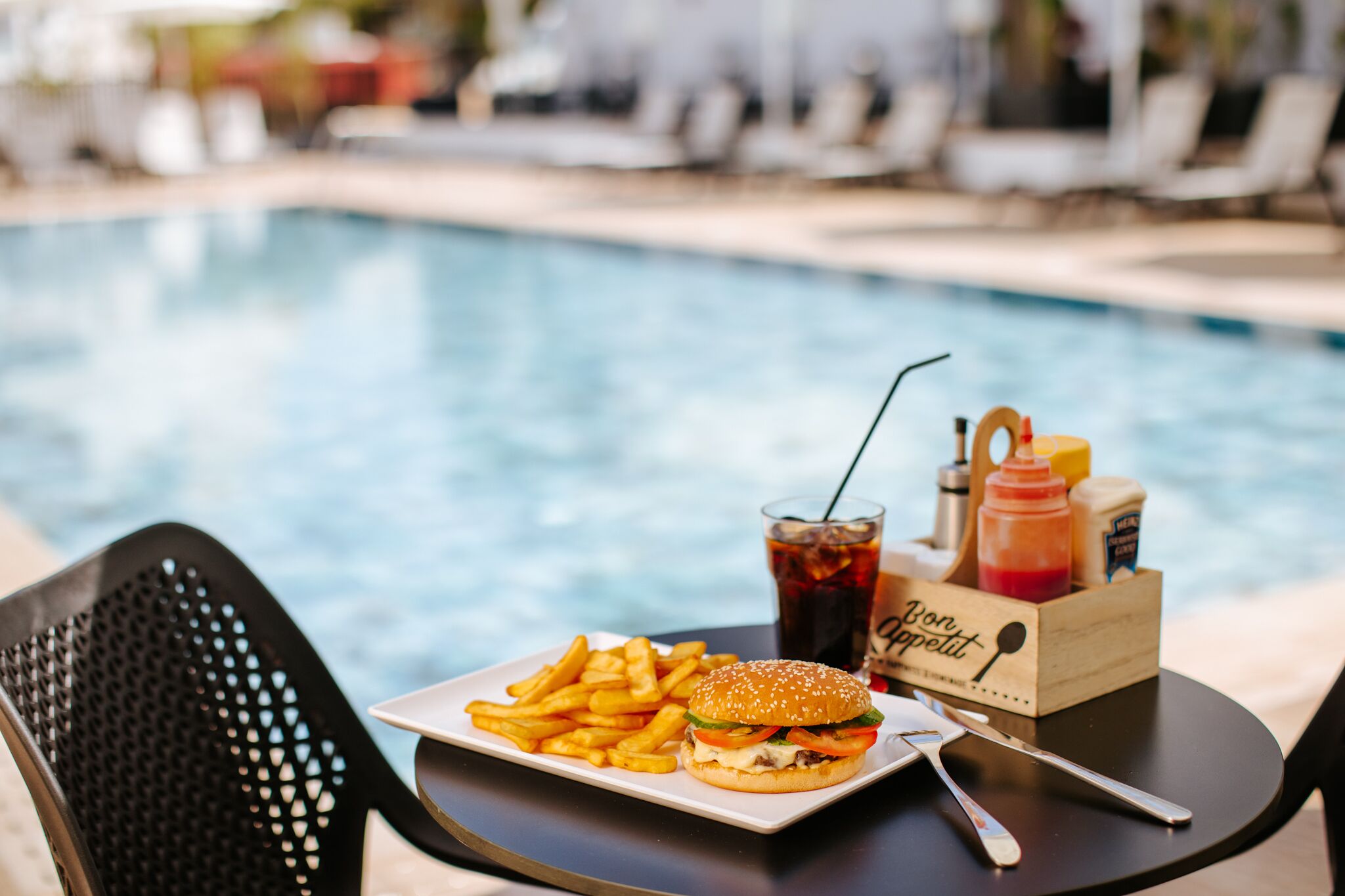 A dining table with a plate of food, a drink, and condiments by a poolside.
