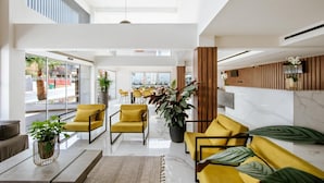 Un hall d’entrée spacieux, avec des fauteuils jaunes, une grande plante en pot et une vue sur l’extérieur.