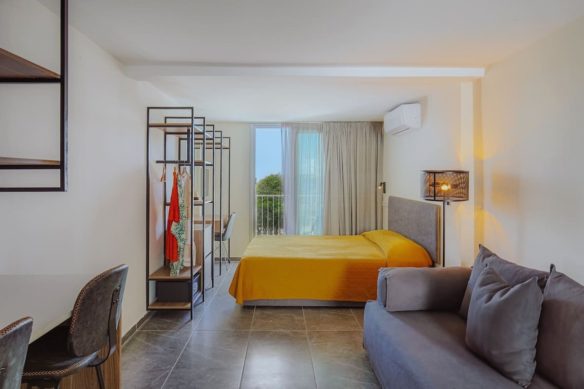 Une chambre d’hôtel avec un lit, un canapé, un bureau et une chaise. Il y a une fenêtre avec des rideaux et un appareil de climatisation fixé au mur.