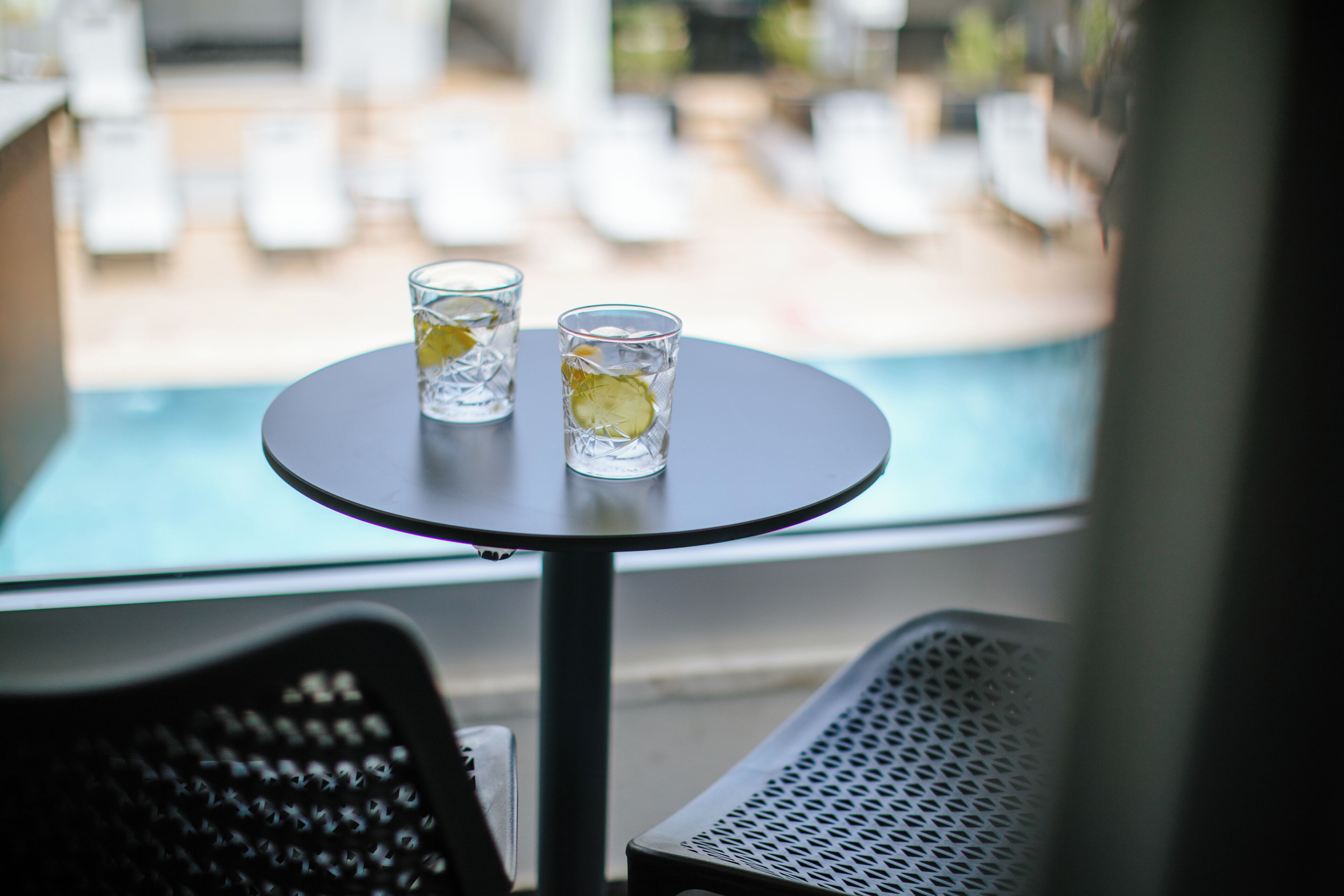 A round table with two glasses of water and a slice of lime. The table is positioned near a window overlooking a pool area with lounge chairs.