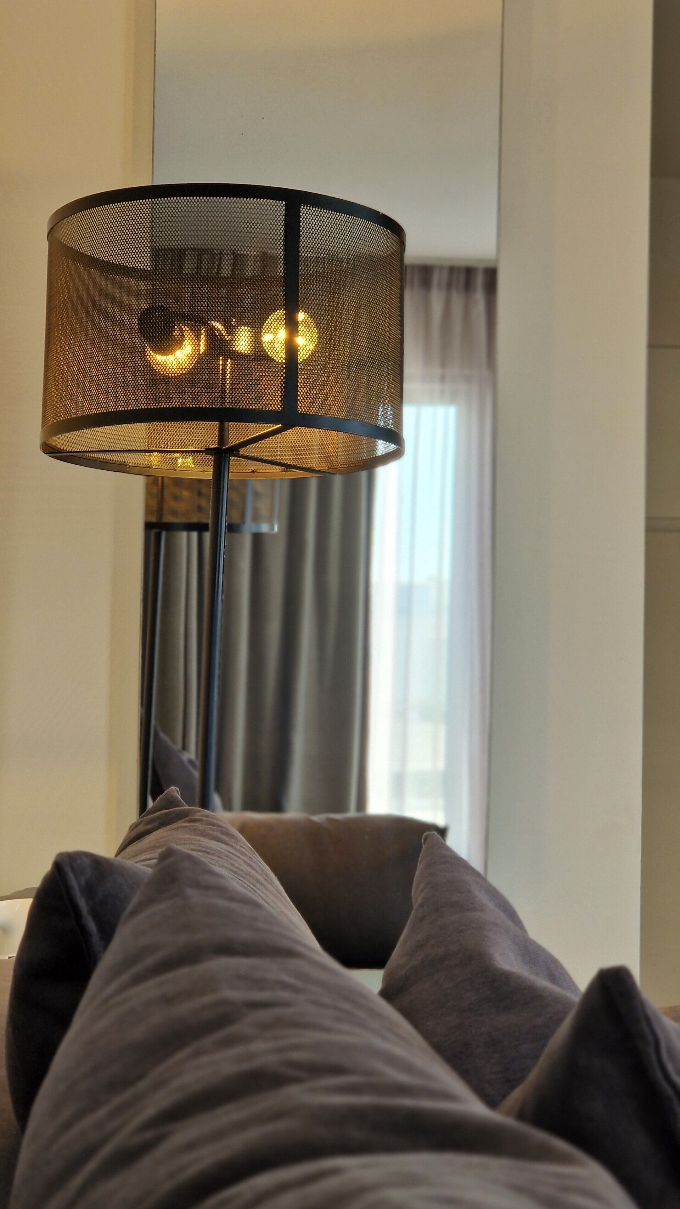 Una habitación con una lámpara de pie grande, una cama con ropa de cama gris y una ventana con cortinas.