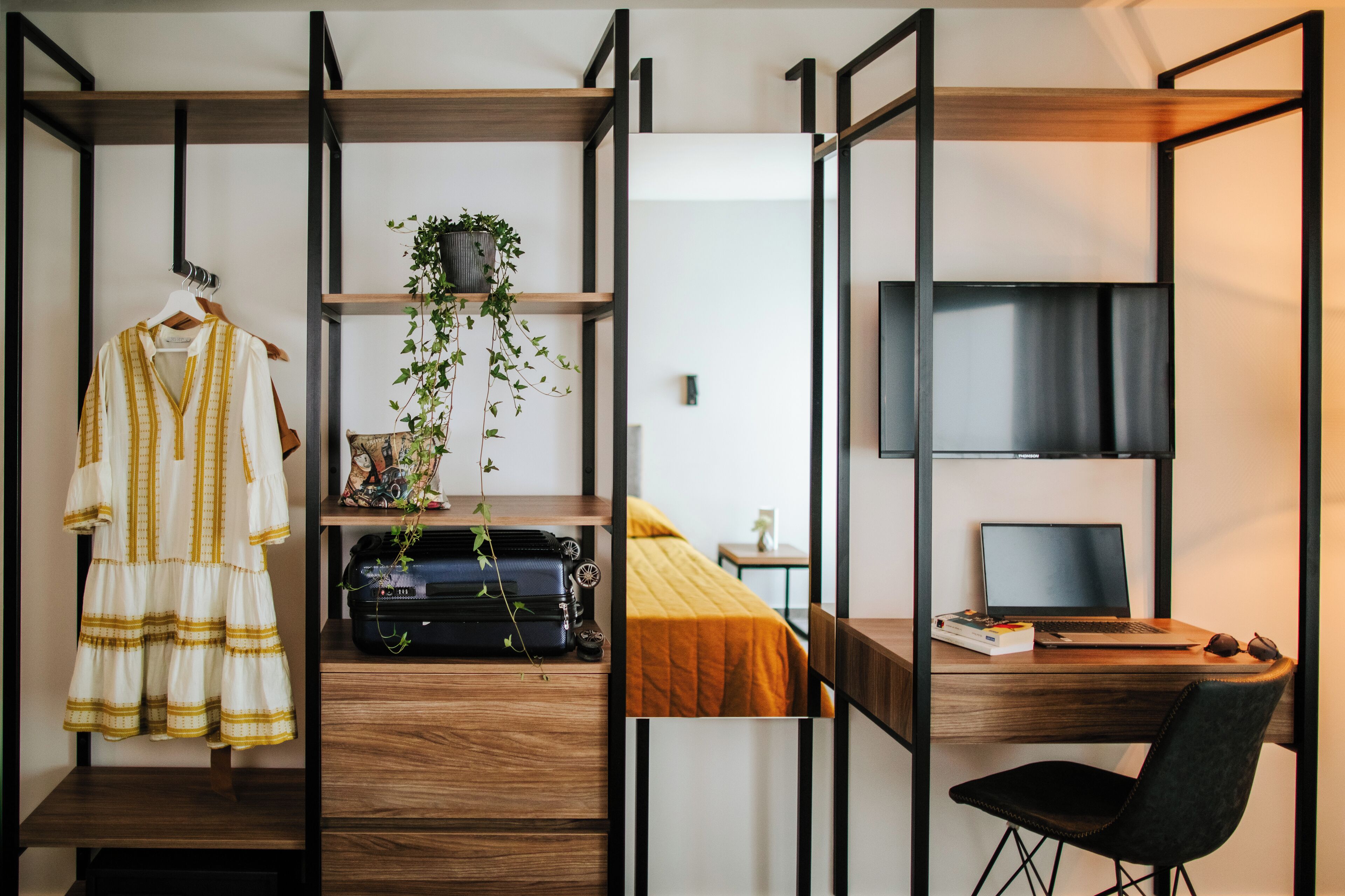 A compact room with a bed, a desk with a laptop, a chair, a hanging plant, and a suitcase.