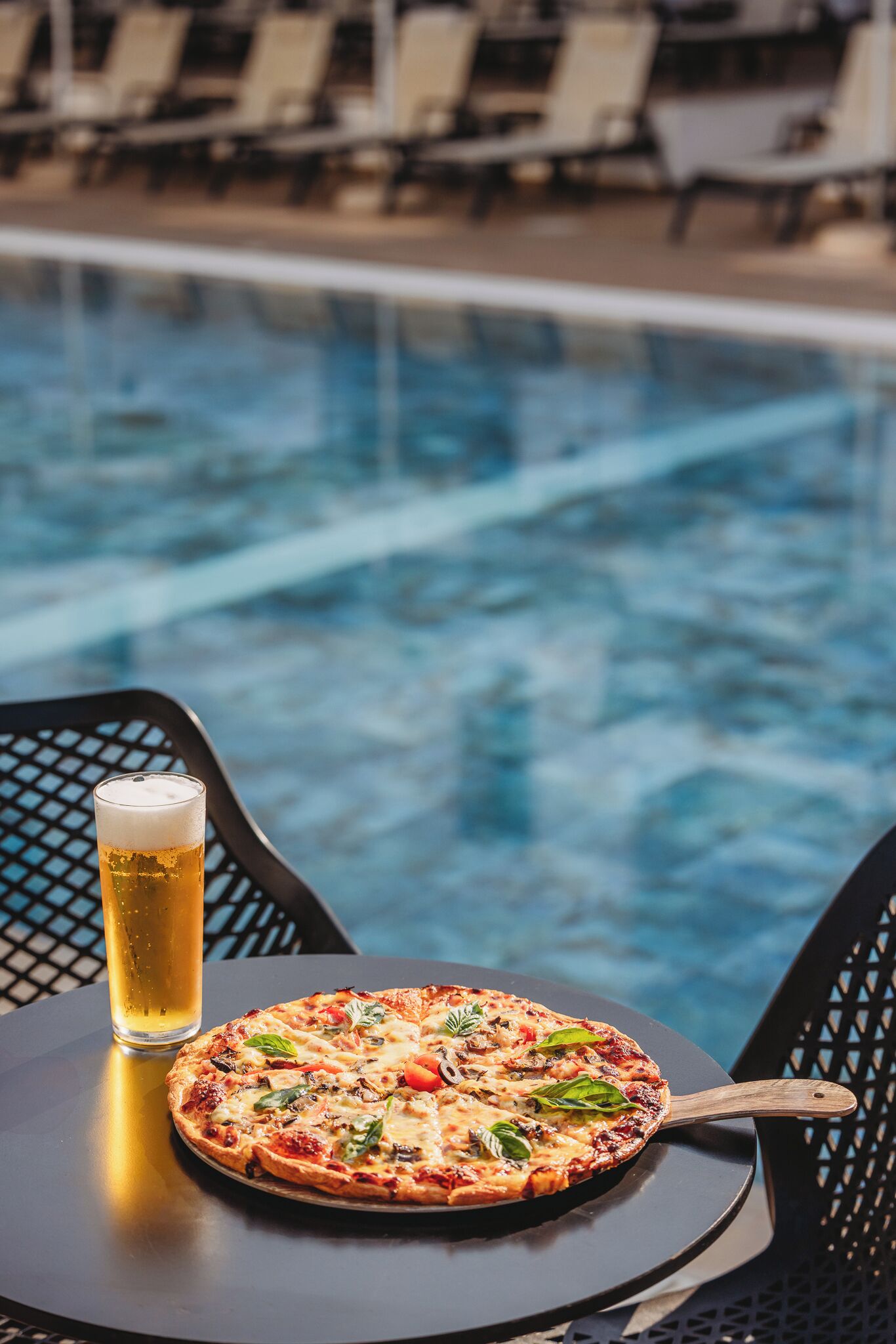 Una pizza y una cerveza sobre una mesa junto a una piscina.