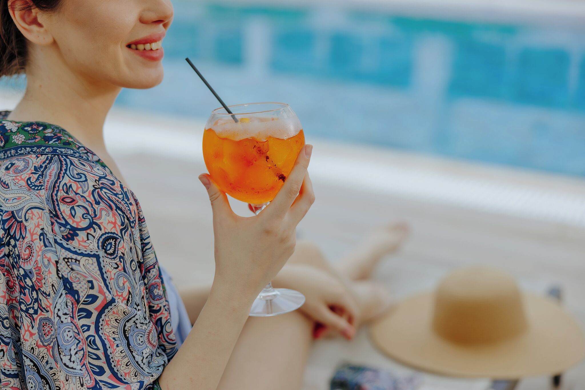 En person holder et cocktailglas med sugerør og sidder ved en pool.