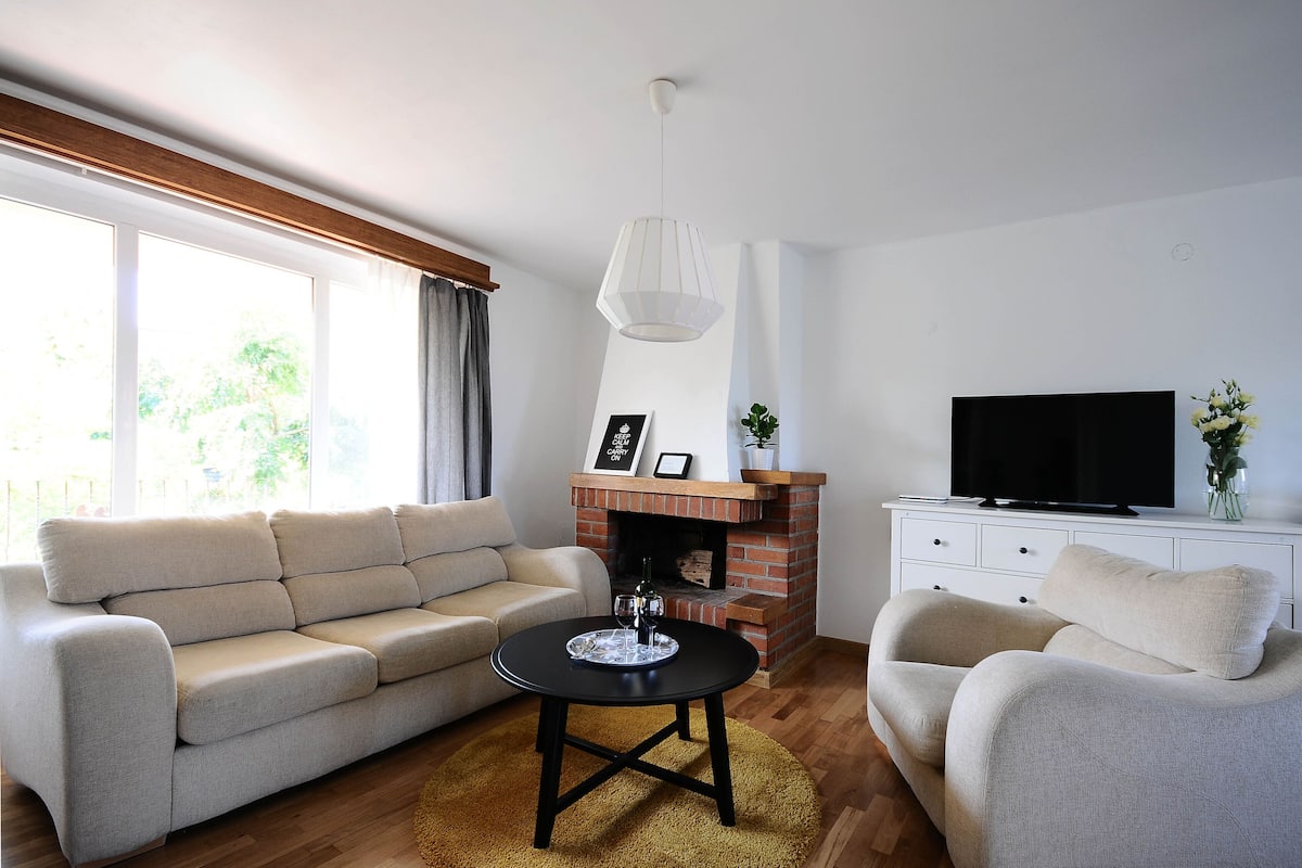 Sala de estar con sofá, sillón y televisor sobre un mueble blanco. Hay una chimenea con repisa y una lámpara colgante.