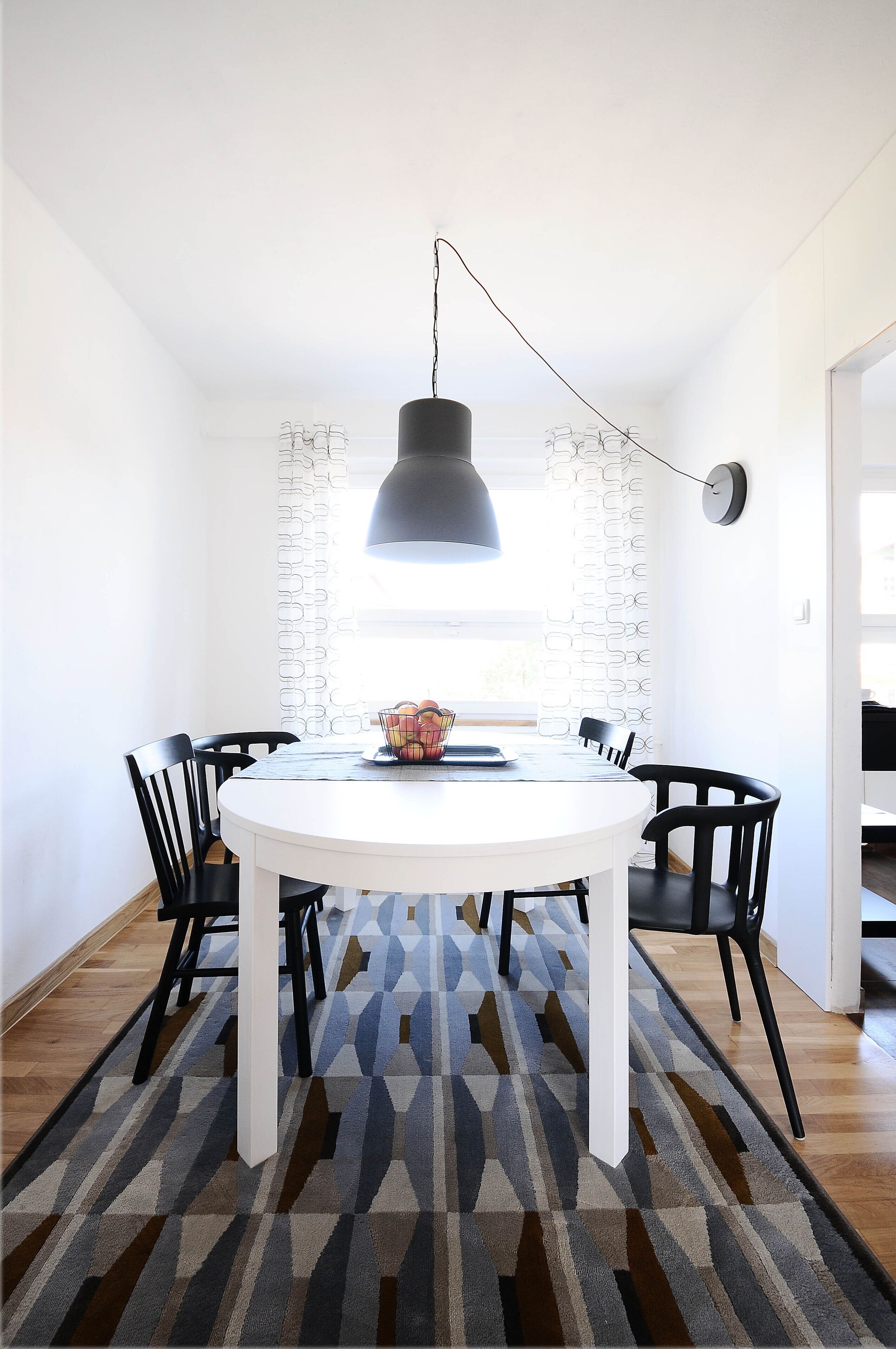 Een eethoek met een ronde tafel, zwarte stoelen, een kamerbreed tapijt met dessin, een hanglamp en een gordijn met patroon.