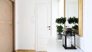 Um corredor com piso de madeira, porta branca, tapete preto, mesa de console branca com espelho e plantas, e uma luminária no teto.