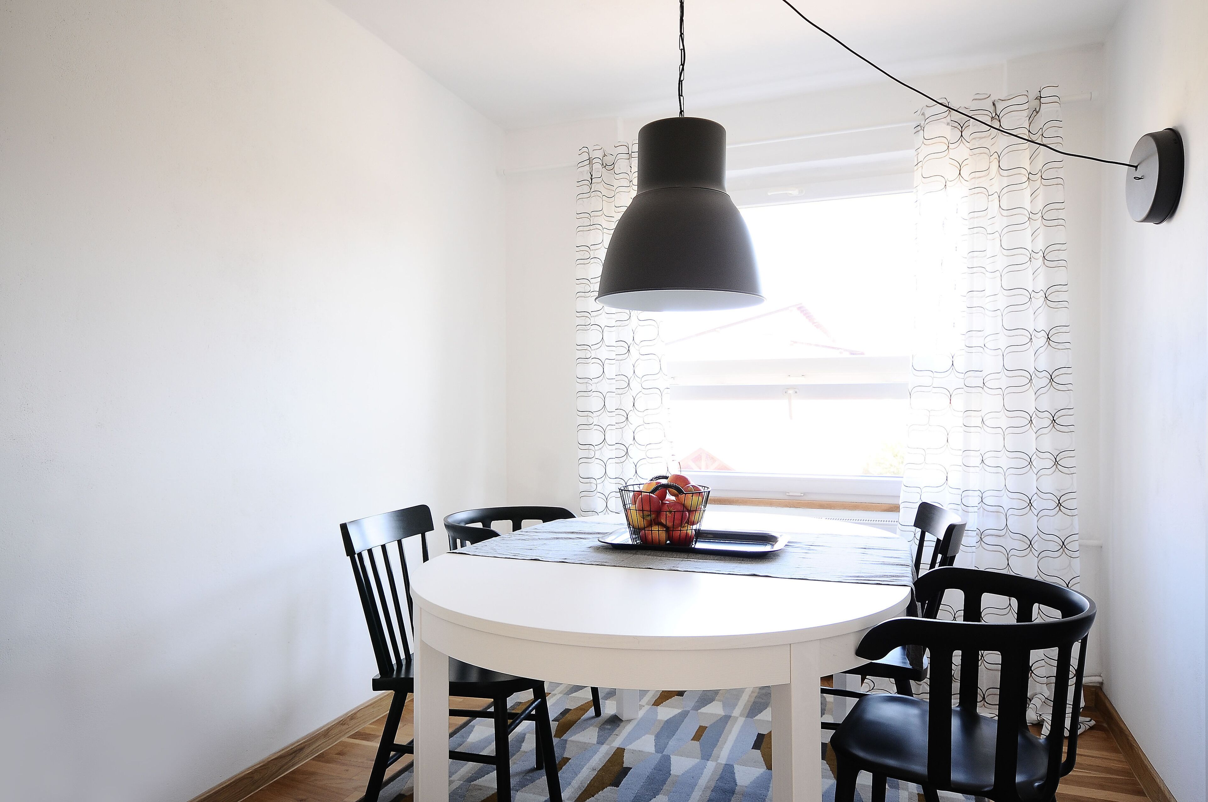 Un coin repas avec une table blanche, des chaises noires, un luminaire suspendu noir et un rideau à motifs.