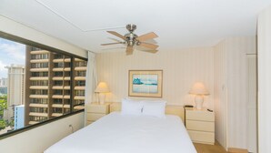 Hotelkamer met een groot bed, twee nachtkastjes met lampen, een plafondventilator, een raam met uitzicht op gebouwen en een lijst met een afbeelding aan de muur.