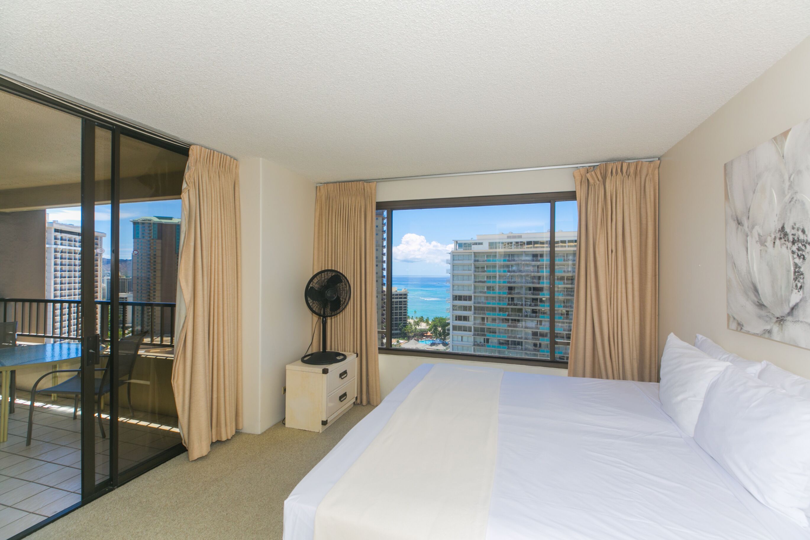 A hotel room with a balcony, a bed, a fan, and a view of the cityscape.