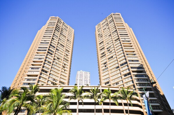 Two tall, beige-colored buildings with multiple balconies, surrounded by palm trees.