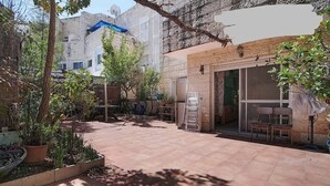Une cour avec un sol carrelé, des plantes en pot, un petit espace aménagé avec des sièges, et un bâtiment doté d’une porte et de fenêtres en verre.