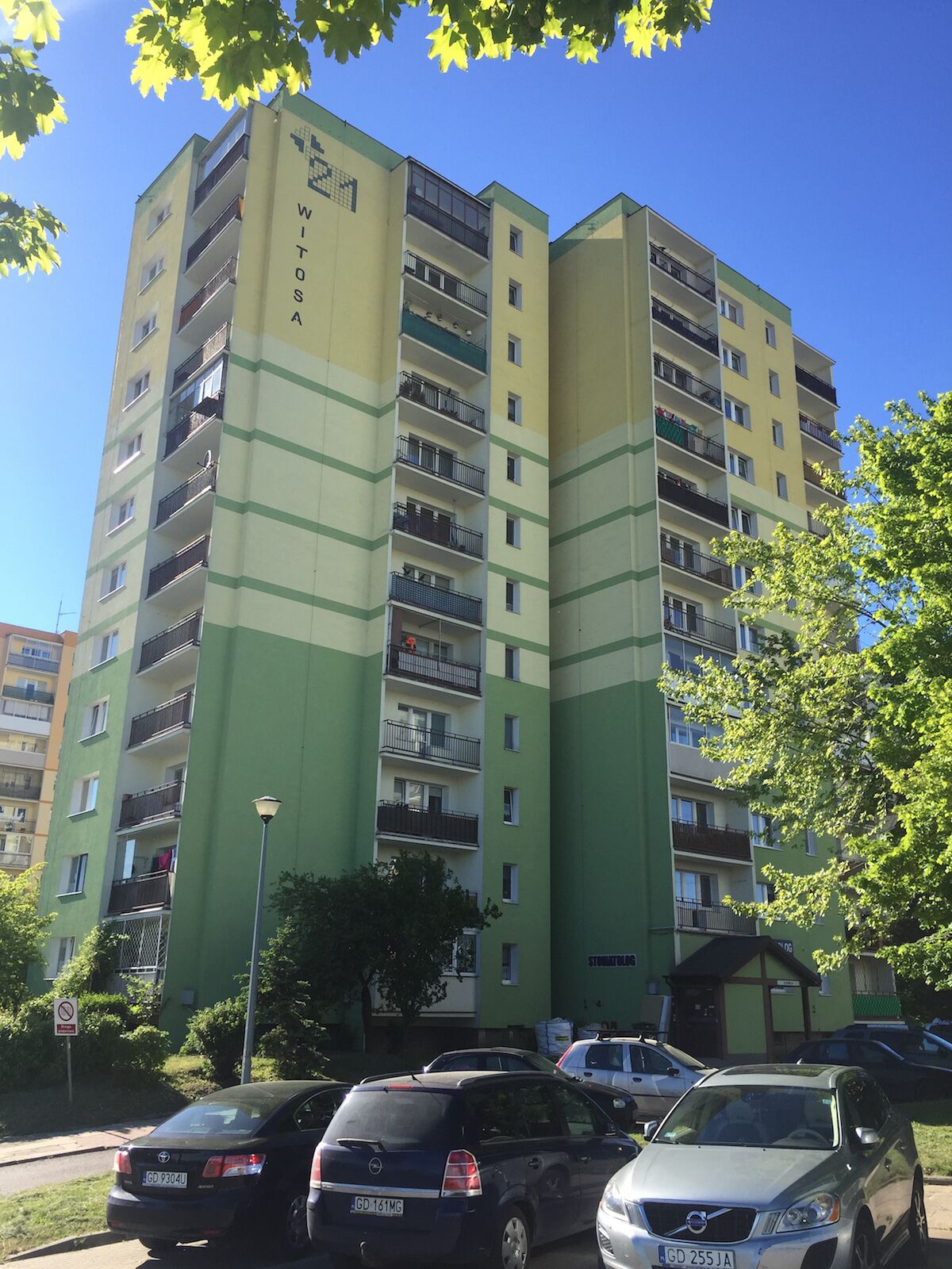 A multi-story residential building with balconies, a parking area with cars, and a clear blue sky.