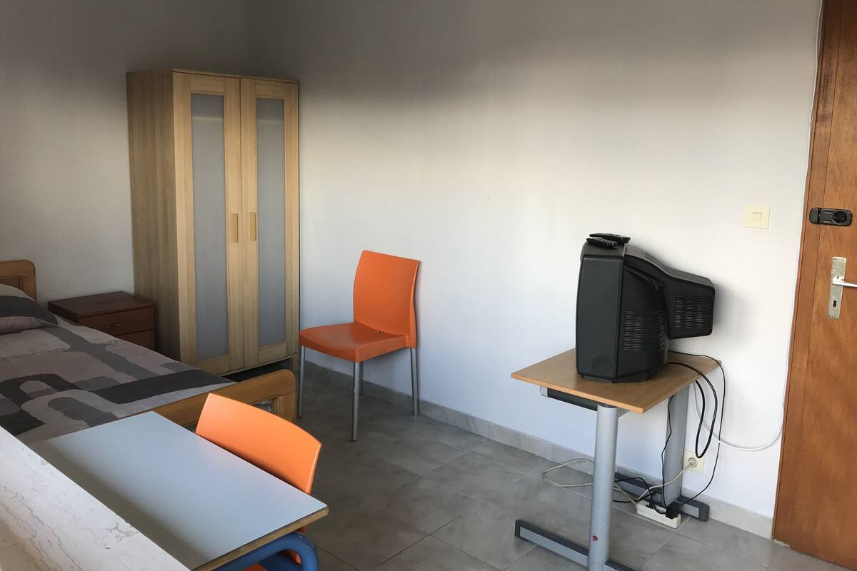 A small, sparsely furnished room with a bed, a wooden wardrobe, a single orange chair, a small table with a television, and a door with a lock.