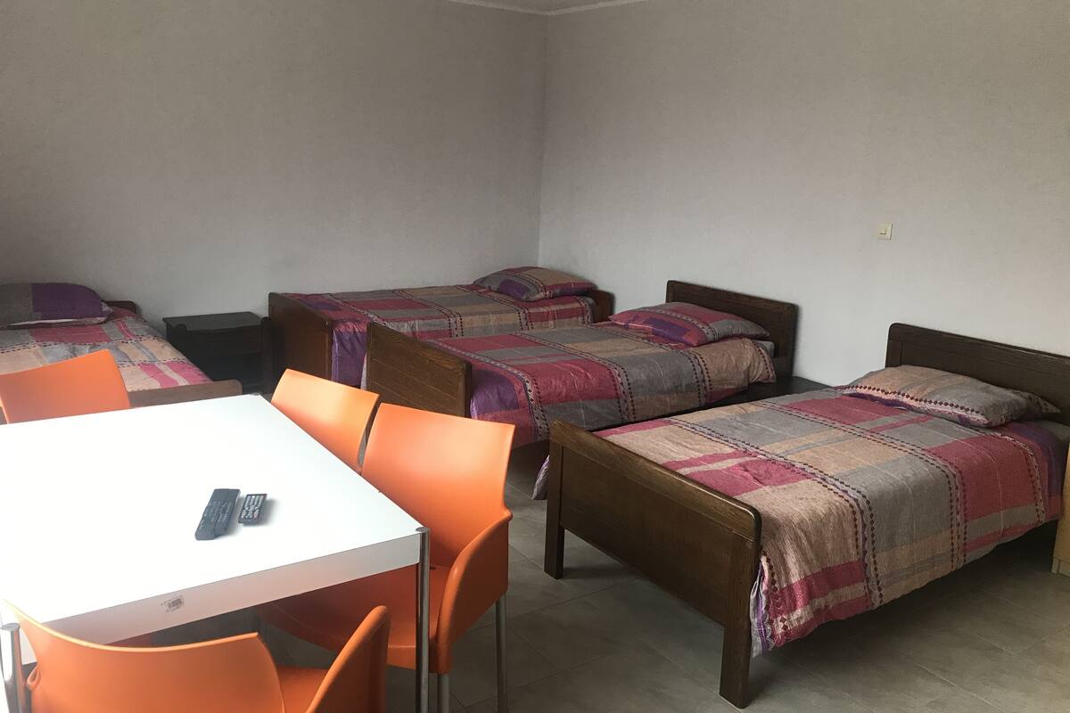 A dormitory room with two single beds, a small table, and two orange chairs.