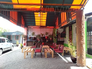 An outdoor seating area with wooden tables and red chairs, surrounded by potted plants and a colorful awning.