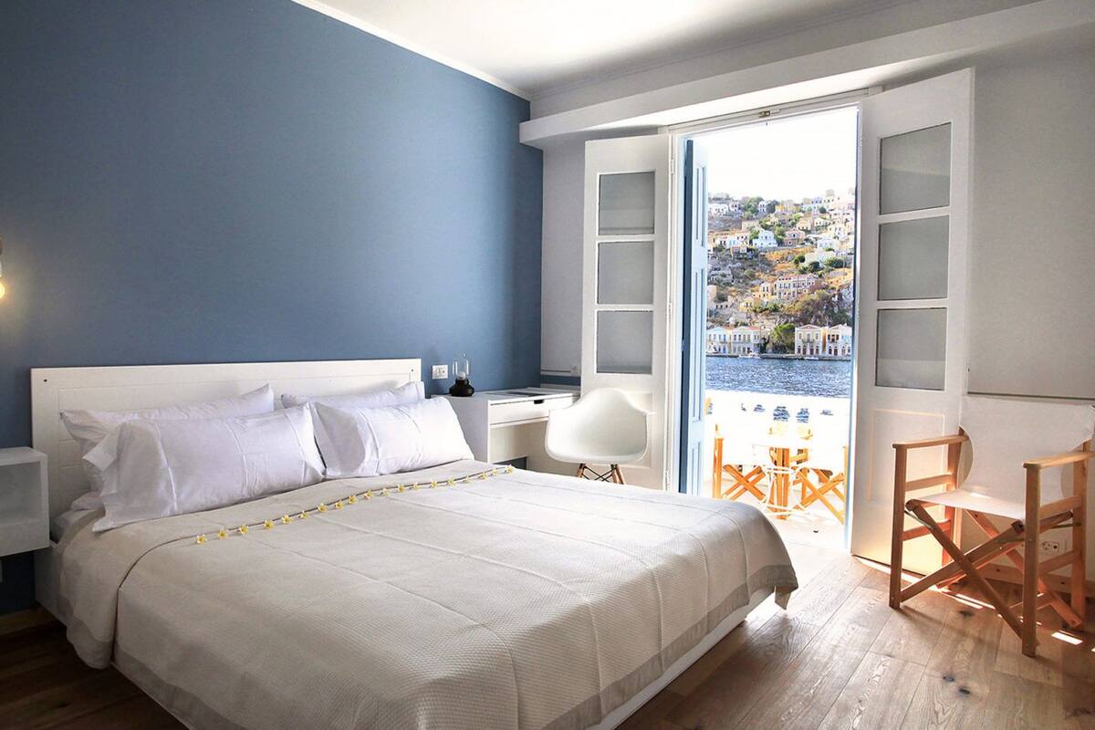 A bedroom with a bed, bedside tables, a chair, and a view of a coastal town through an open door.