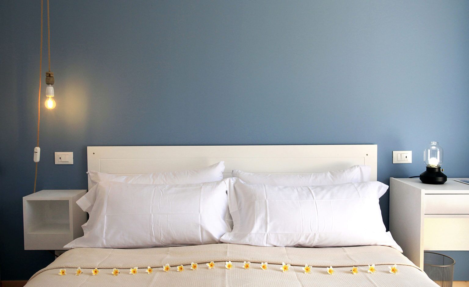 Une chambre à coucher avec un lit, des oreillers blancs, une table de chevet avec une lampe et une prise électrique au mur.