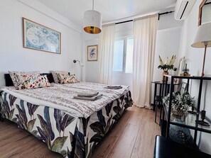 Une chambre à coucher avec un lit, une table de chevet, une lampe, une fenêtre et un mur orné d’un tableau encadré.