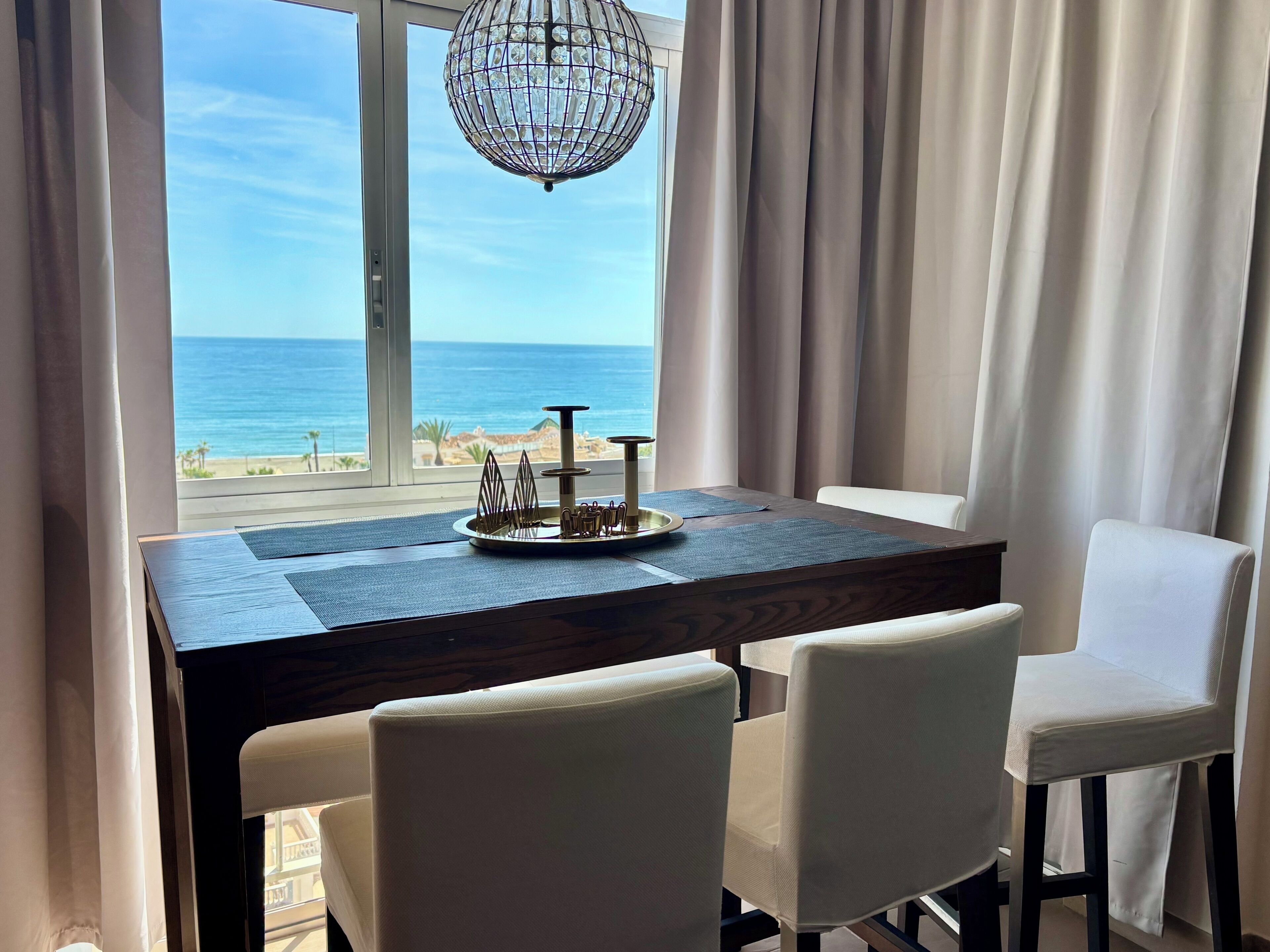 Uma sala de jantar com vista para a praia, um candelabro e uma mesa com cadeiras.