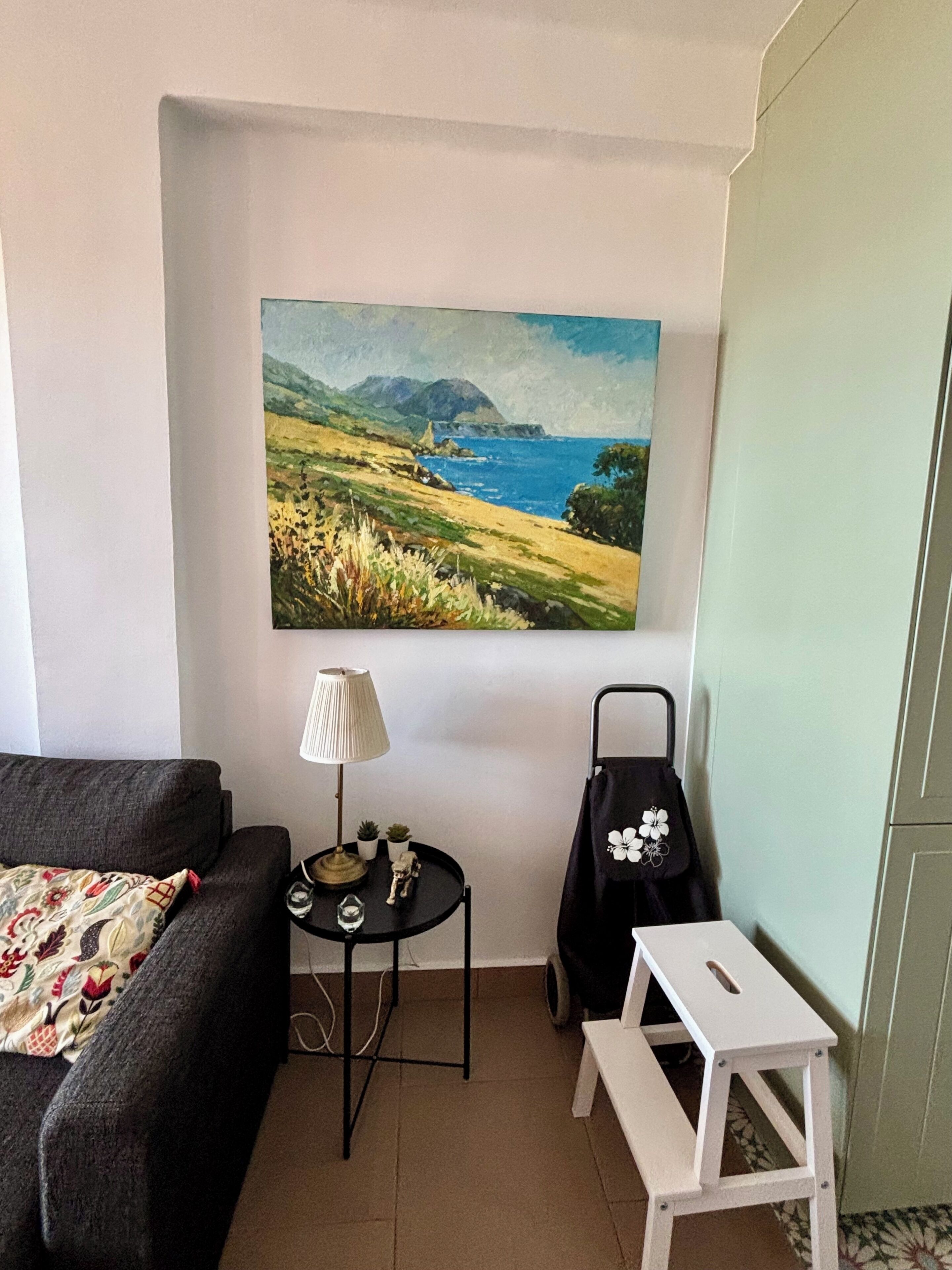 A room with a sofa, a small table with a lamp, a painting of a landscape, and a white bench.