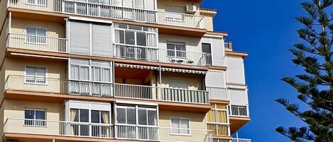 Un edificio residencial de varios pisos con balcones, unidades de aire acondicionado y un cielo azul despejado al fondo.