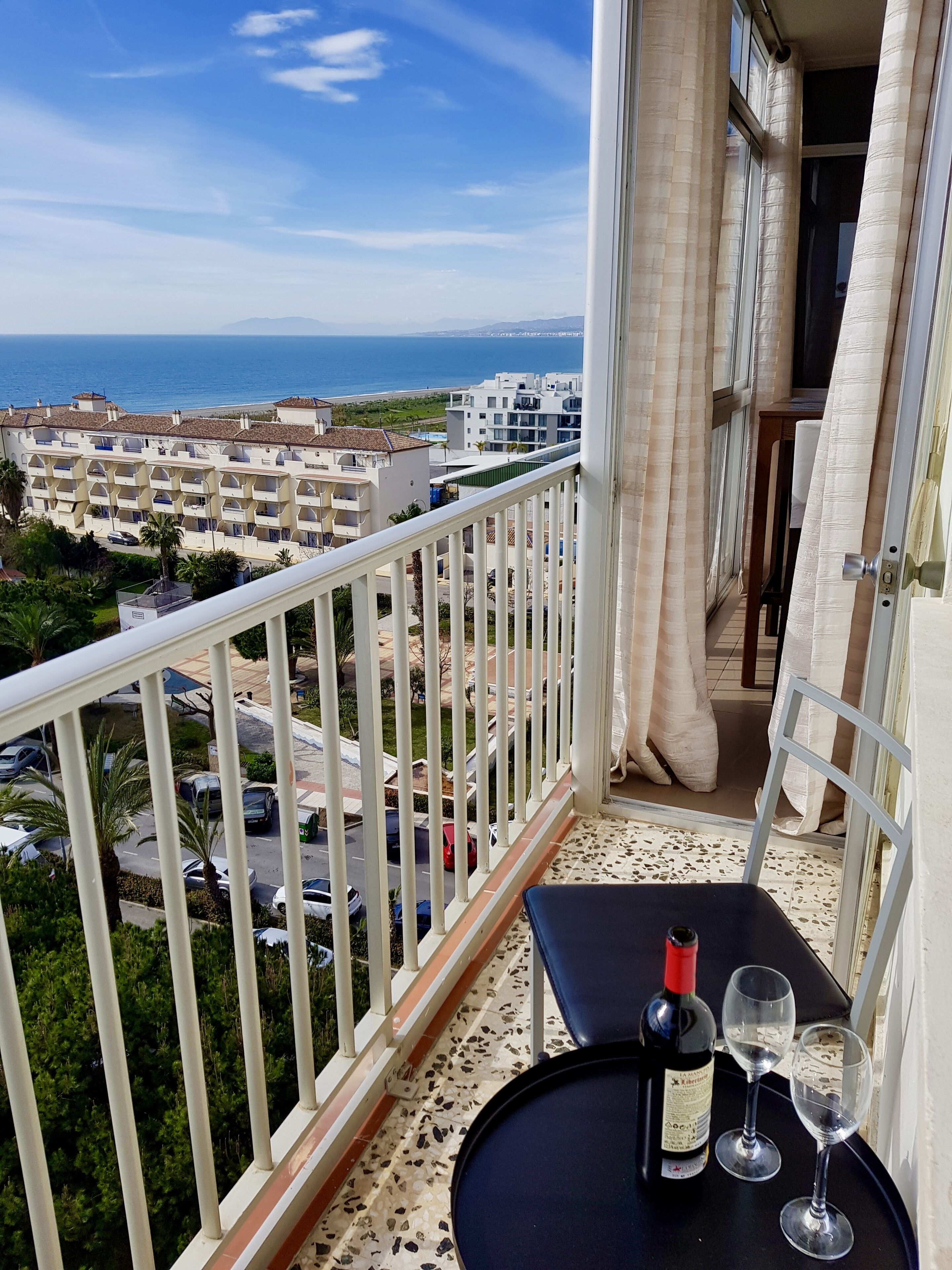 Un balcón con una mesa, una botella y dos vasos. El balcón da a una zona residencial y al mar.