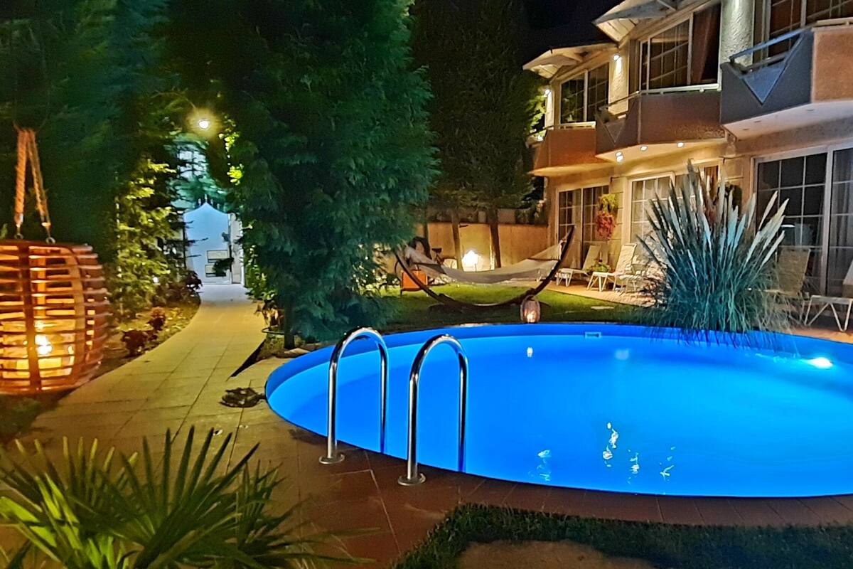 Ein Swimmingpool mit Metallleiter, umgeben von einer Terrasse mit Stühlen und einer Hängematte.