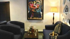 A living room with a dark blue sofa, a small coffee table with bottles and glasses, and a painting on the wall. - De Brass Suites Hotel (Yenagoa)