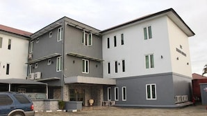 A multi-story building with a grey and white facade, a covered parking area, and a car parked in front. - De Brass Suites Hotel (Yenagoa)